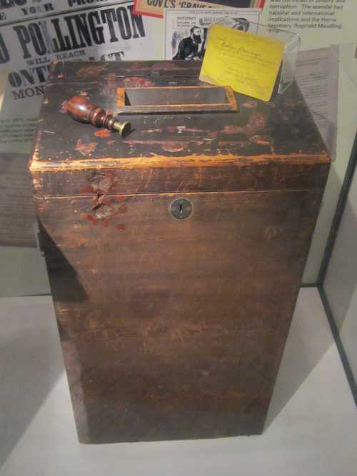 A weathered wooden ballot box with a keyhole, accompanied by a small note and a wooden mallet, displayed in a glass case.