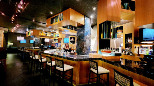 A modern bar with wooden accents, colorful decor, and a variety of drinks displayed on shelves.