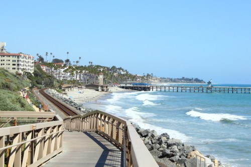 Scenic coastal view with a boardwalk, beach, palm trees, and a pier under a clear blue sky.