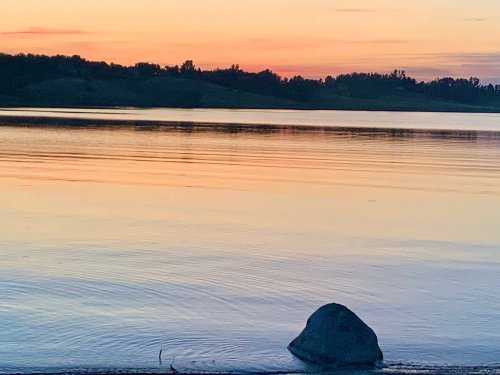 A tranquil lake at sunset, with soft colors reflecting on the water and a large rock in the foreground.