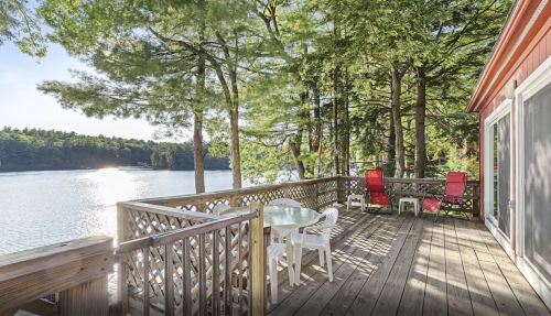A serene lakeside deck surrounded by trees, featuring a table and chairs, with sunlight reflecting on the water.
