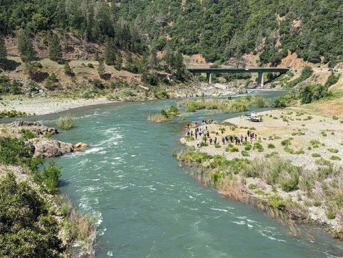 A winding river flows through a green landscape, with a group of people gathered on the shore near a bridge.