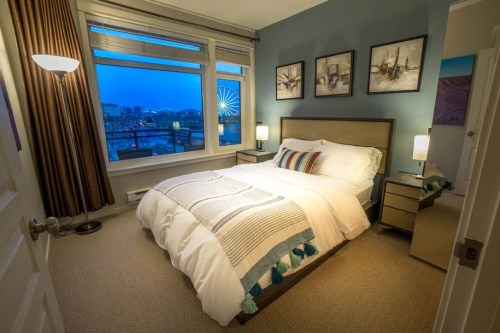 Cozy bedroom with a large window showcasing a city view, featuring a bed, nightstands, and decorative artwork.