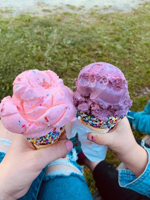 Two hands holding colorful ice cream cones, one pink with sprinkles and one purple, against a grassy background.