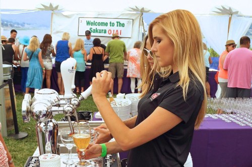 A woman pours a drink from a tap at a festival, with a crowd enjoying the event in the background.