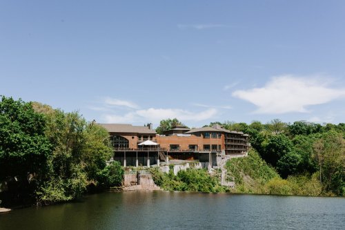A modern building on a hillside overlooking a calm lake, surrounded by lush greenery and a clear blue sky.
