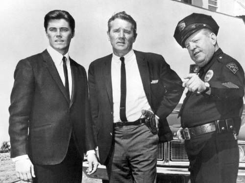 Three men in suits, one in a police uniform, stand together outdoors near a police car, discussing something.