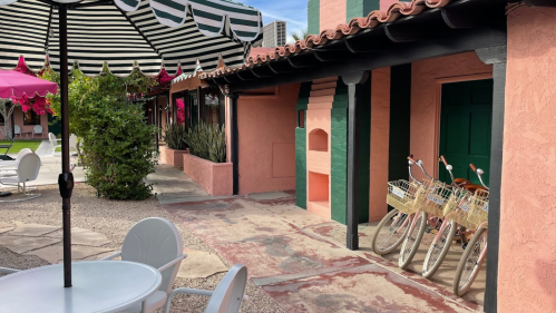 A colorful outdoor area with pink walls, green doors, and bicycles parked near a patio with white tables and chairs.