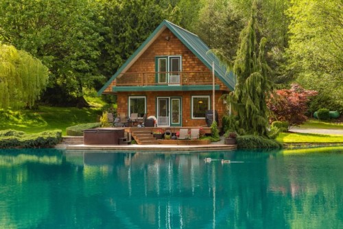 A charming wooden cabin by a serene turquoise lake, surrounded by lush greenery and trees. Ducks swim nearby.