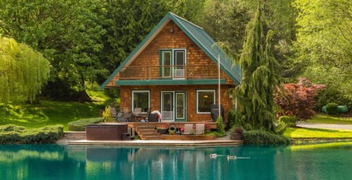 A charming wooden cabin by a serene pond, surrounded by lush greenery and a hot tub on the deck.
