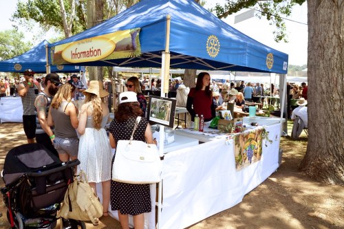 A busy outdoor event with a blue information tent, people gathering around, and trees in the background.