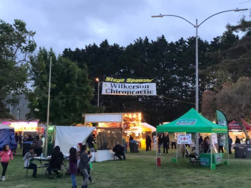 A lively outdoor event with food stalls, tents, and a banner for Wilkerson Chiropractic in the background.