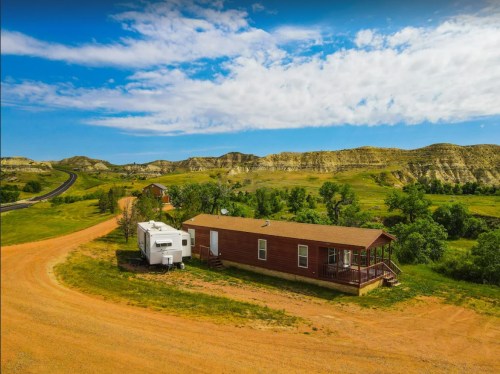 A scenic view of a red house and RV on a dirt road, surrounded by green fields and rolling hills under a blue sky.