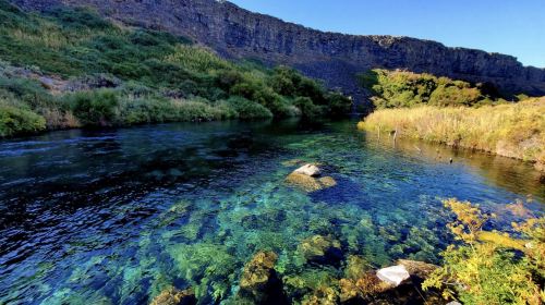 A serene river flows through a lush landscape, with clear water revealing rocks beneath the surface and steep cliffs in the background.