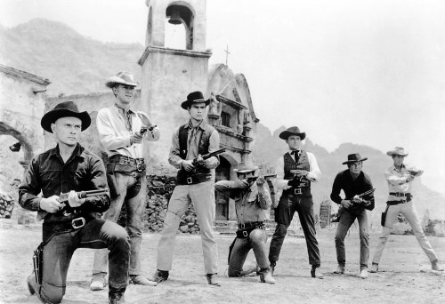 A group of seven cowboys in Western attire, posing with guns in a desert landscape near an old building.