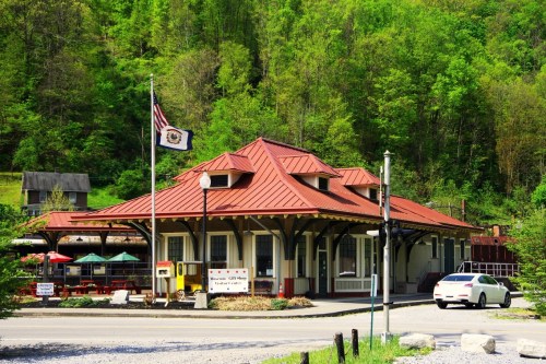 Historic train station with a red roof, surrounded by greenery and outdoor seating, featuring an American flag.