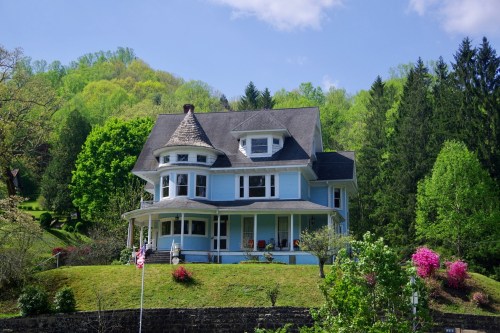 A large blue Victorian house with a turret, surrounded by lush green trees and colorful flowers on a sunny day.