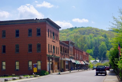 A quiet street lined with brick buildings, green hills in the background, and a blue sky with fluffy clouds.