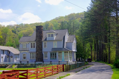 A large, light blue Victorian-style house with a stone chimney, surrounded by trees and a red wooden fence.