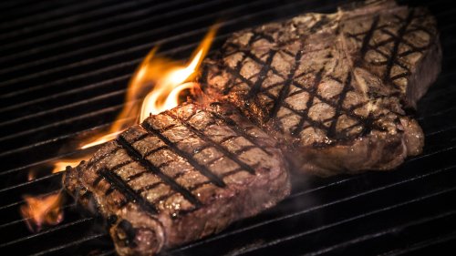 Two steaks grilling on a barbecue, with flames and grill marks visible.