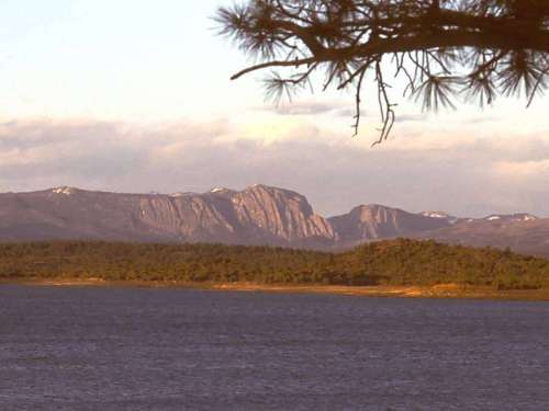 A scenic view of mountains rising above a calm lake, with trees framing the foreground and a cloudy sky above.