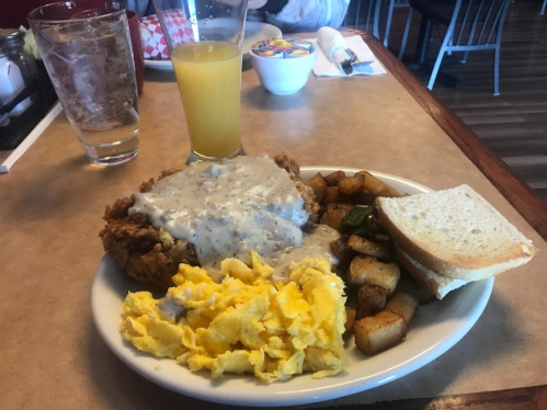 A plate of fried chicken with gravy, scrambled eggs, seasoned potatoes, and a slice of bread, alongside a glass of orange juice.