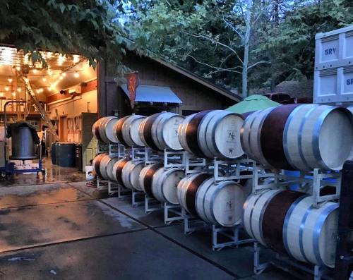 A row of wine barrels outside a rustic winery, with warm lights glowing from the building in the background.