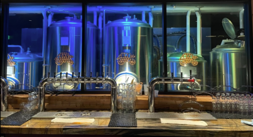 A brewery bar with shiny taps and large brewing tanks illuminated by blue lights in the background.