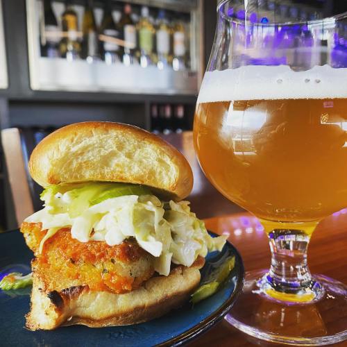 A crispy chicken sandwich with lettuce on a bun, served with a glass of golden beer in a cozy restaurant setting.