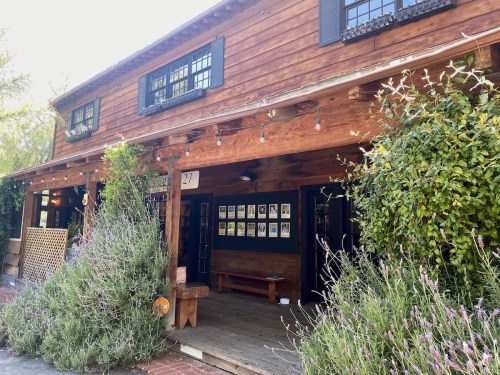 A rustic wooden building with a porch, surrounded by greenery and featuring a display of framed photos on the wall.