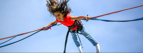 A girl in a red shirt joyfully jumps on a bungee trampoline against a clear blue sky.