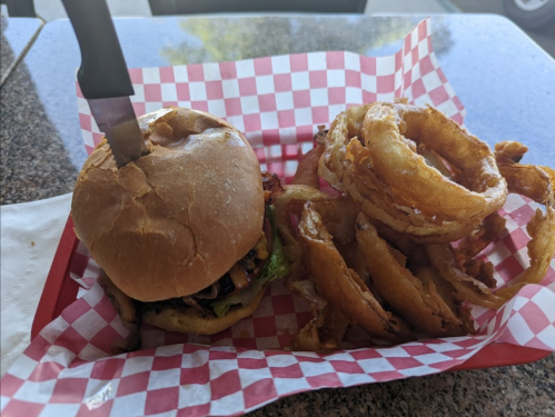 A burger with toppings and a knife on top, served with a side of crispy onion rings in a red-checkered basket.