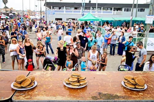 A crowded outdoor event with people enjoying the festivities, featuring plates of burgers in the foreground.