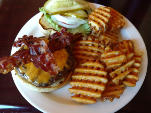 A plate with a cheeseburger topped with bacon, lettuce, pickles, and onions, served with waffle fries.