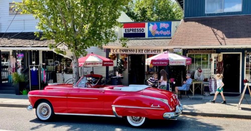 A red convertible parked in front of an ice cream shop with outdoor seating and people enjoying their time.