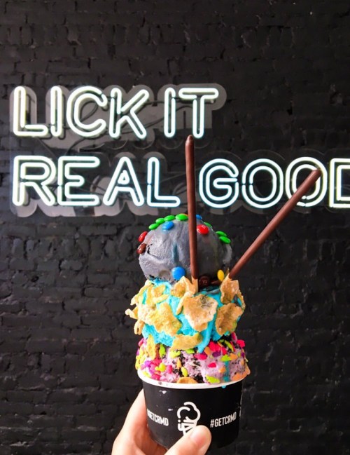 A colorful ice cream cone topped with sprinkles and chocolate sticks, in front of a neon sign that says "LICK IT REAL GOOD."