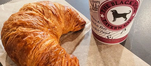 A golden croissant next to a coffee cup from The Black Dog Tavern, resting on a piece of parchment paper.