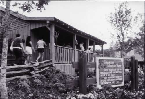 Black and white photo of a wooden building with people walking up, surrounded by trees and flowers. Sign visible in front.