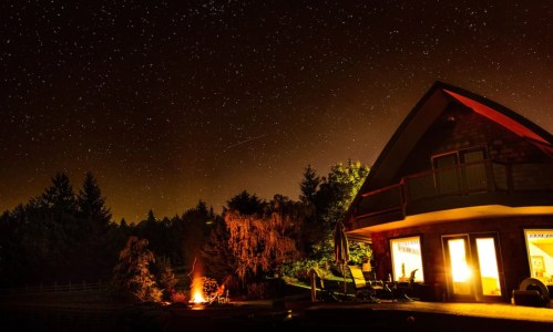 A cozy cabin under a starry night sky, with a warm glow from the windows and a campfire nearby.