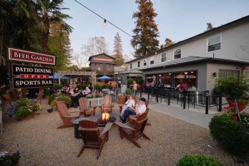 A cozy outdoor beer garden with a fire pit, surrounded by plants, featuring patio dining and a sports bar.