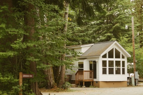 A cozy white house with a large front porch, surrounded by tall green trees, near a sign for Cross Road.