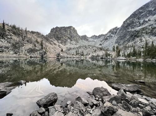 A serene mountain landscape reflected in calm water, surrounded by rocky terrain and sparse trees.