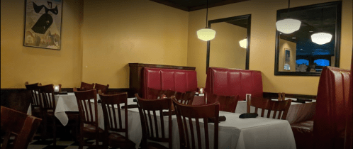 Cozy restaurant interior with red booths, wooden chairs, and white tablecloths, featuring soft lighting and a mirror.