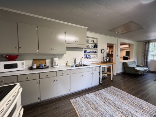 A modern kitchen with gray cabinets, a white countertop, and a cozy seating area with a rug and natural light.