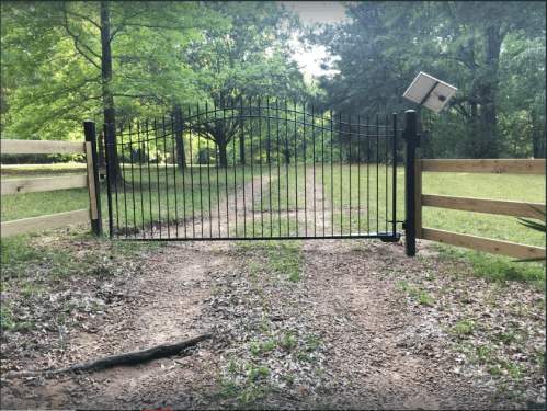 A black metal gate opens to a dirt path lined by trees and wooden fences in a green, serene landscape.