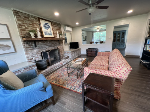 Cozy living room with a brick fireplace, patterned sofa, blue armchair, and wooden furniture. Natural light from windows.