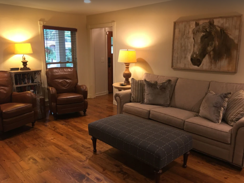 Cozy living room with a beige sofa, two brown armchairs, a blue ottoman, and warm lighting. Horse painting on the wall.