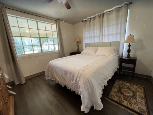 Cozy bedroom with a white bed, wooden nightstand, lamps, and large window letting in natural light.
