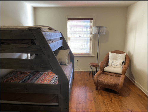 A cozy bedroom featuring a wooden bunk bed, a chair with a cushion, and a window with blinds.