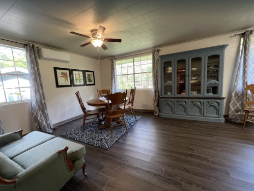 Cozy dining area with a round table, wooden chairs, a blue cabinet, and large windows letting in natural light.